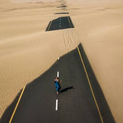 A Lone Woman Walking On A Sand Covered Road In Dubai's Desert, Captured From Above.