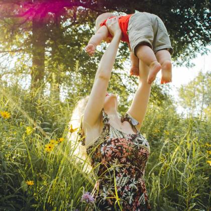 A Joyful Mother Lifts Her Baby In A Sunny Summer Meadow, Capturing A Moment Of Love And Happiness.