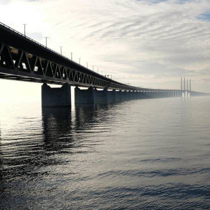A Stunning View Of A Large Steel Bridge Reflecting On The Tranquil Water Under A Cloudy Sky.