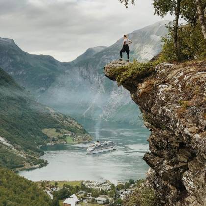 Person Enjoys Breathtaking View From A Cliff Above Geiranger Fjord, Norway, With Clear Skies And Cruise Ship Below.