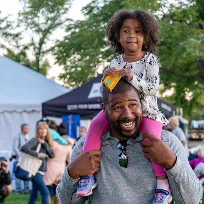 A Joyful Father Carrying His Daughter At An Outdoor Festival Surrounded By People And Tents.