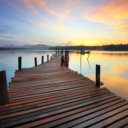 A Tranquil Wooden Jetty Stretching Into A Calm Lake At Sunrise With Colorful Sky Reflections.