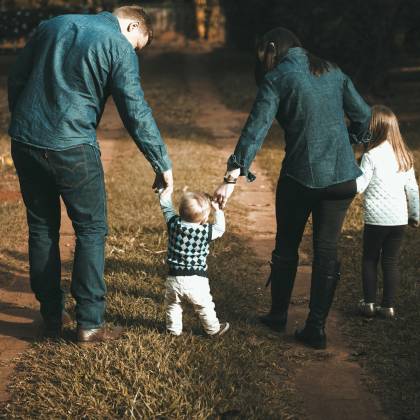 A Family Of Four Walks Hand In Hand On A Path, Enjoying A Sunny Day Outdoors.