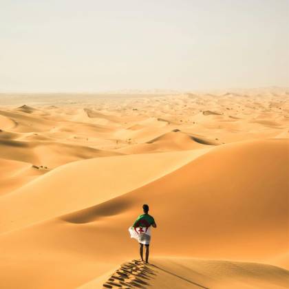 A Solitary Traveler Walks Across The Vast Sand Dunes Of The Sahara Desert In Algeria.