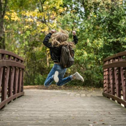 A Blonde Girl Jumps Exuberantly On A Wooden Bridge Surrounded By Autumn Foliage, Capturing The Essence Of Joy And Freedom.