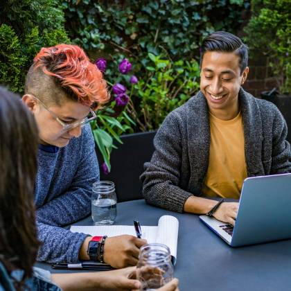 Young Professionals Working Together On A Project Outdoors With Laptops And Notebooks In A Vibrant Garden Setting.