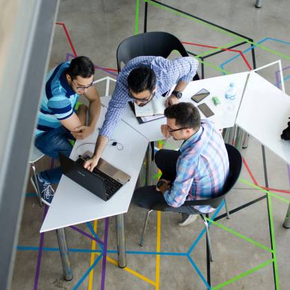 Three Men Collaborating Over A Laptop In A Modern, Geometric Themed Office Space.