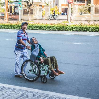 A Caring Man Aids An Elderly Woman In A Wheelchair Across A Sunny Urban Street In Cần Thơ, Vietnam.