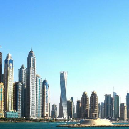 Panoramic View Of Dubai's Towering Skyscrapers Set Against A Vibrant Blue Sky And Waterfront.
