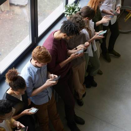 A Group Of Diverse Office Workers Standing By A Window Using Smartphones, Capturing Modern Workplace Connectivity.