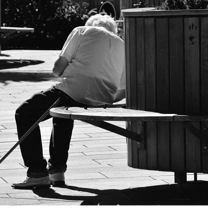 A Black And White Image Of A Senior Man Sitting On A Bench With A Cane In An Urban Park.