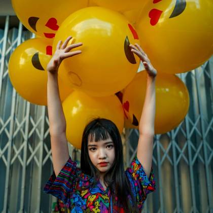A Young Woman Poses With Bright Emoji Balloons Against An Urban Background.