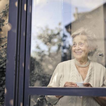 Elderly Woman Smiles By A Glass Window, Holding A Cup, In A Peaceful Setting.