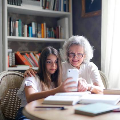 Elderly Woman And Girl Happily Taking A Selfie On A Smartphone In A Cozy Library Setting.