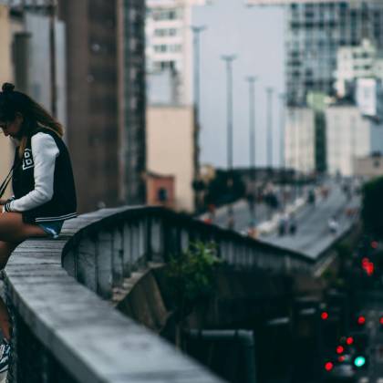 A Woman Casually Sitting On A Bridge In A Bustling Cityscape During The Day.