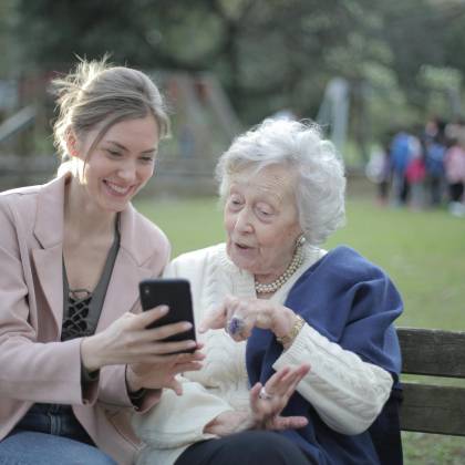 Delighted Female Relatives Sitting Together On Wooden Bench In Park And Browsing Mobile Phone While Learning Using