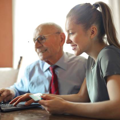 Young Woman In Casual Clothes Helping Senior Man In Formal Shirt With Paying Credit Card In Internet Using Laptop While Sitting At Table
