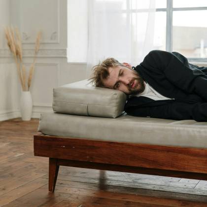 A Bearded Man Lying On A Couch In A Bright, Minimalistic Room, Looking Contemplative.