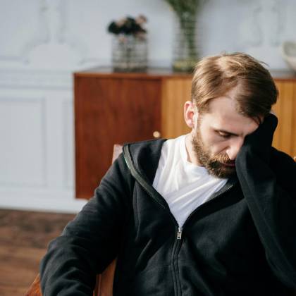 Bearded Man In A Black Hoodie Sitting In A Chair Indoors, Appearing Deep In Thought.