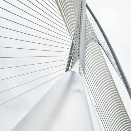 Low Angle View Of A Modern Suspension Bridge With Steel Cables And Architectural Lines Under A Cloudy Sky.