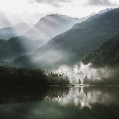 Breathtaking View Of A Foggy Mountain Landscape With Sun Rays Piercing Through, Reflecting On A Serene Lake.