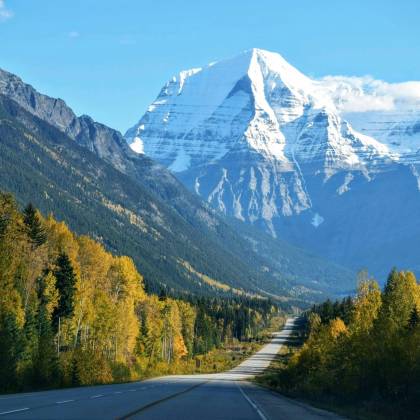 Stunning Autumn Scene With A Road Leading To A Snow Capped Mountain Under A Clear Blue Sky.