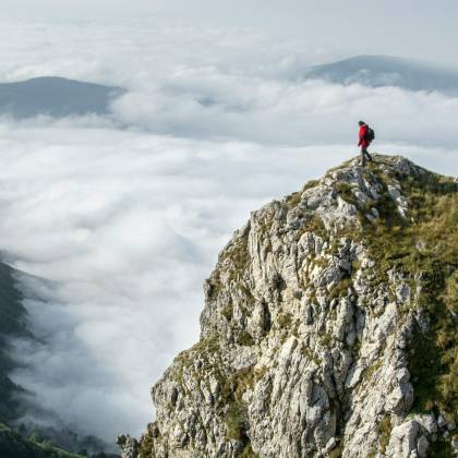 Hiker In Red Standing On A Rocky Mountain Summit Enveloped By Clouds.