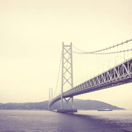 A Stunning View Of A Suspension Bridge Spanning The Sea With A Hazy Backdrop.