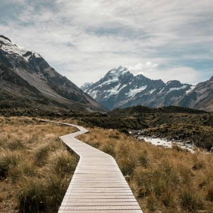 Wooden Walkway Leads Through Rugged Landscape Towards Majestic Snow Capped Mountains.
