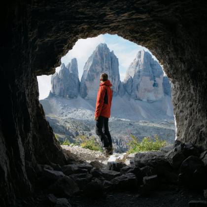 Man In Red Jacket Standing In Cave Entrance, Viewing Tre Cime Di Lavaredo In Italy.
